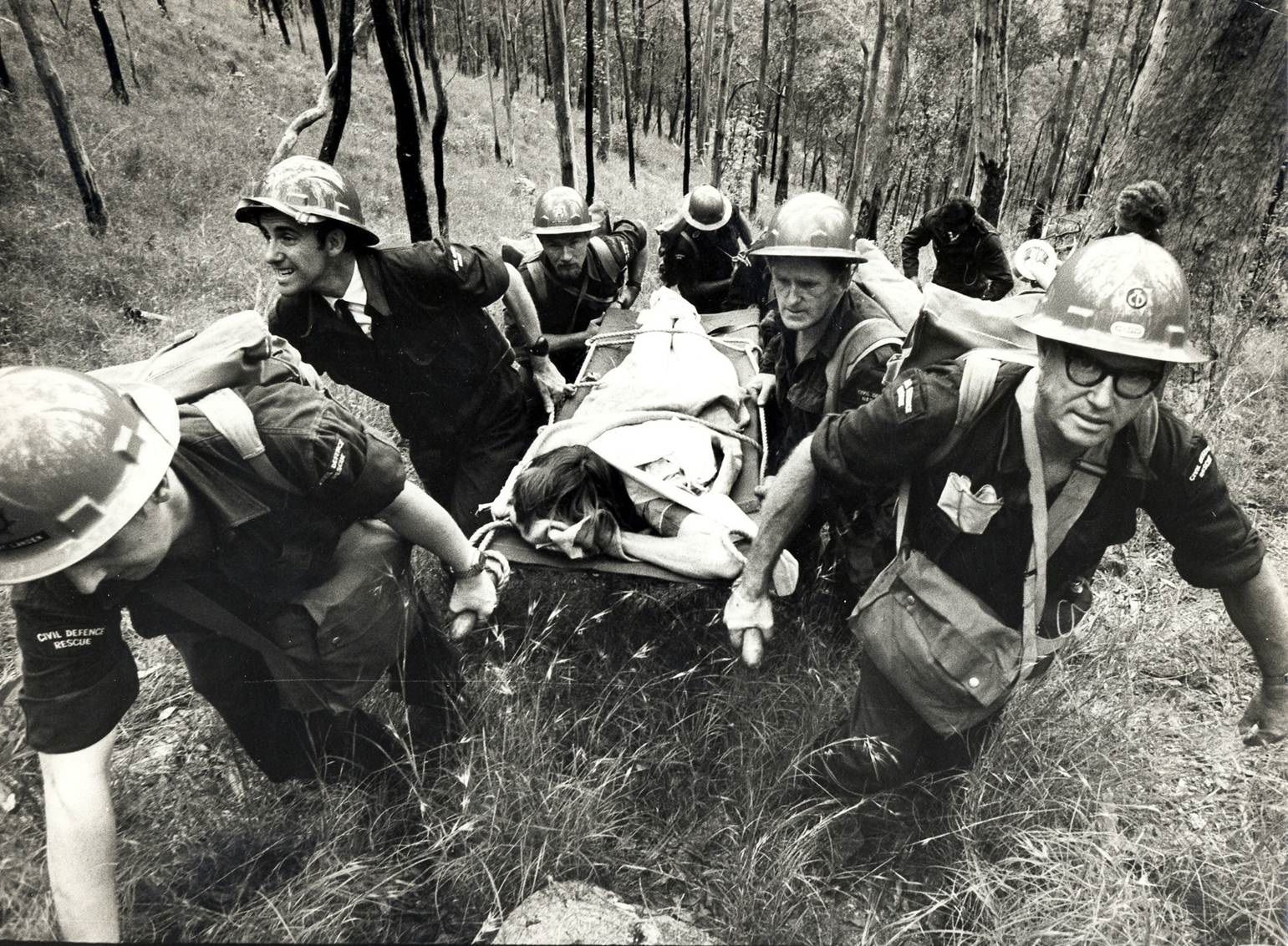 Black and white image of four men in civil defence uniforms carrying person in stretcher up a hill in the bush.