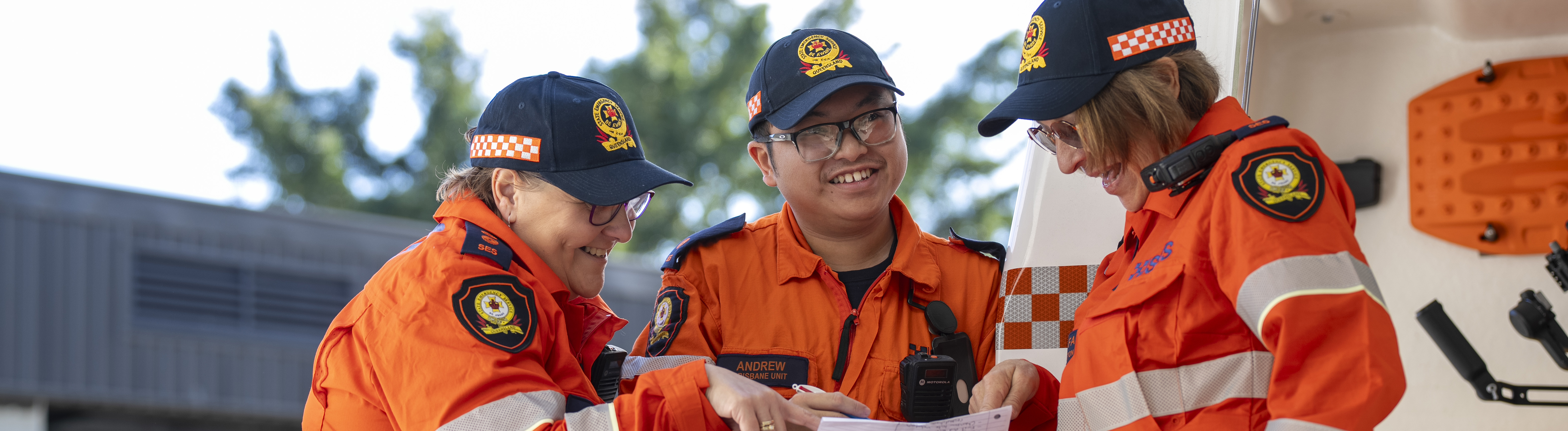 Image of three SES members looking up observing and discussing notes on paper. All in orange uniform and hats. Standing in front of SES vehicle.