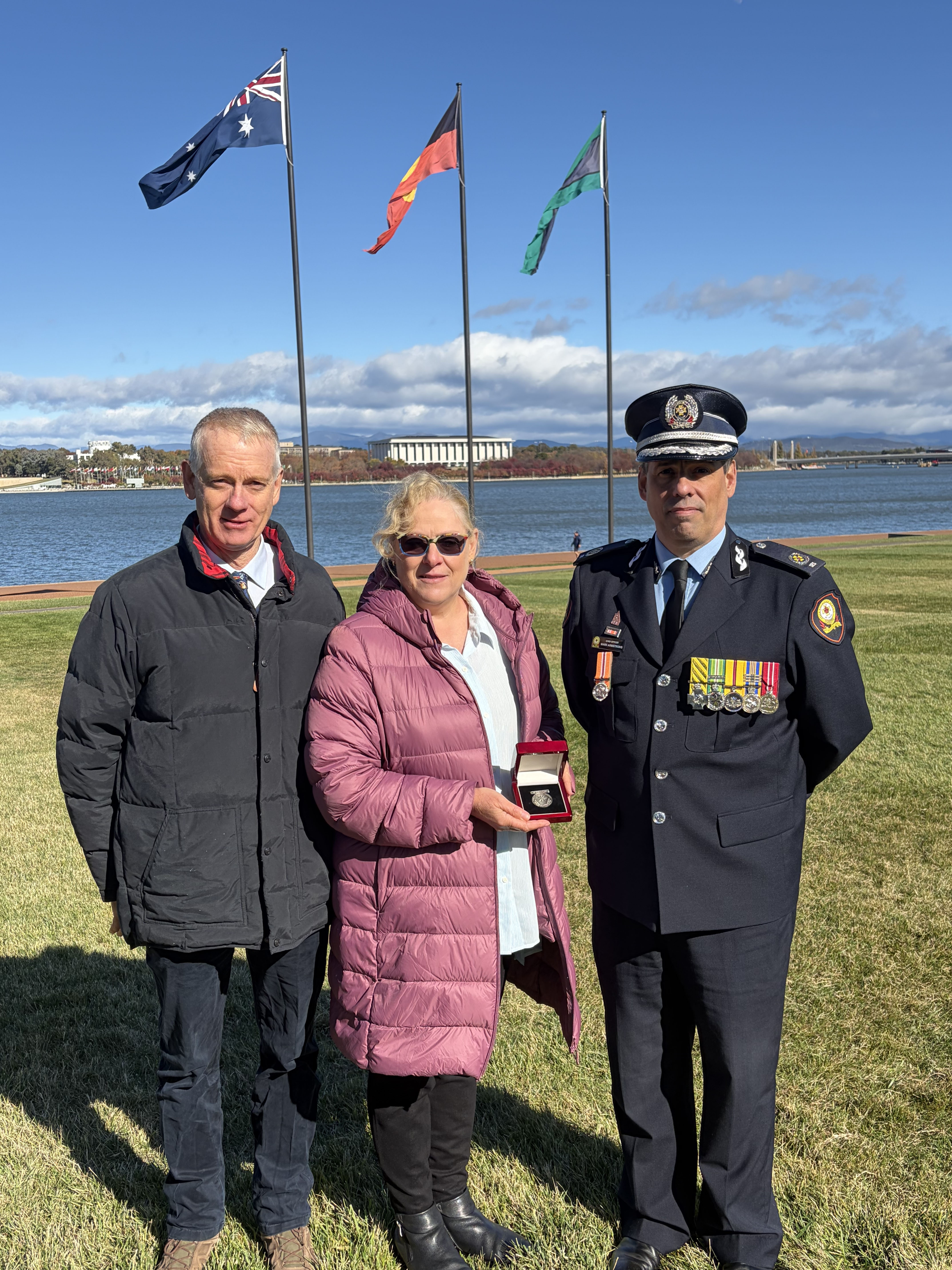 Rowen Taylor, Melody Taylor and Chief Officer Mark Armstrong CSC stand in front of Lake Burly Griffin and the Australian, Aboriginal and Torres Strait Islander flags at the National Emergency Services Memorial.