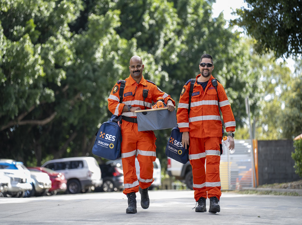 Two SES personnel in orange safety uniforms carrying equipment bags.