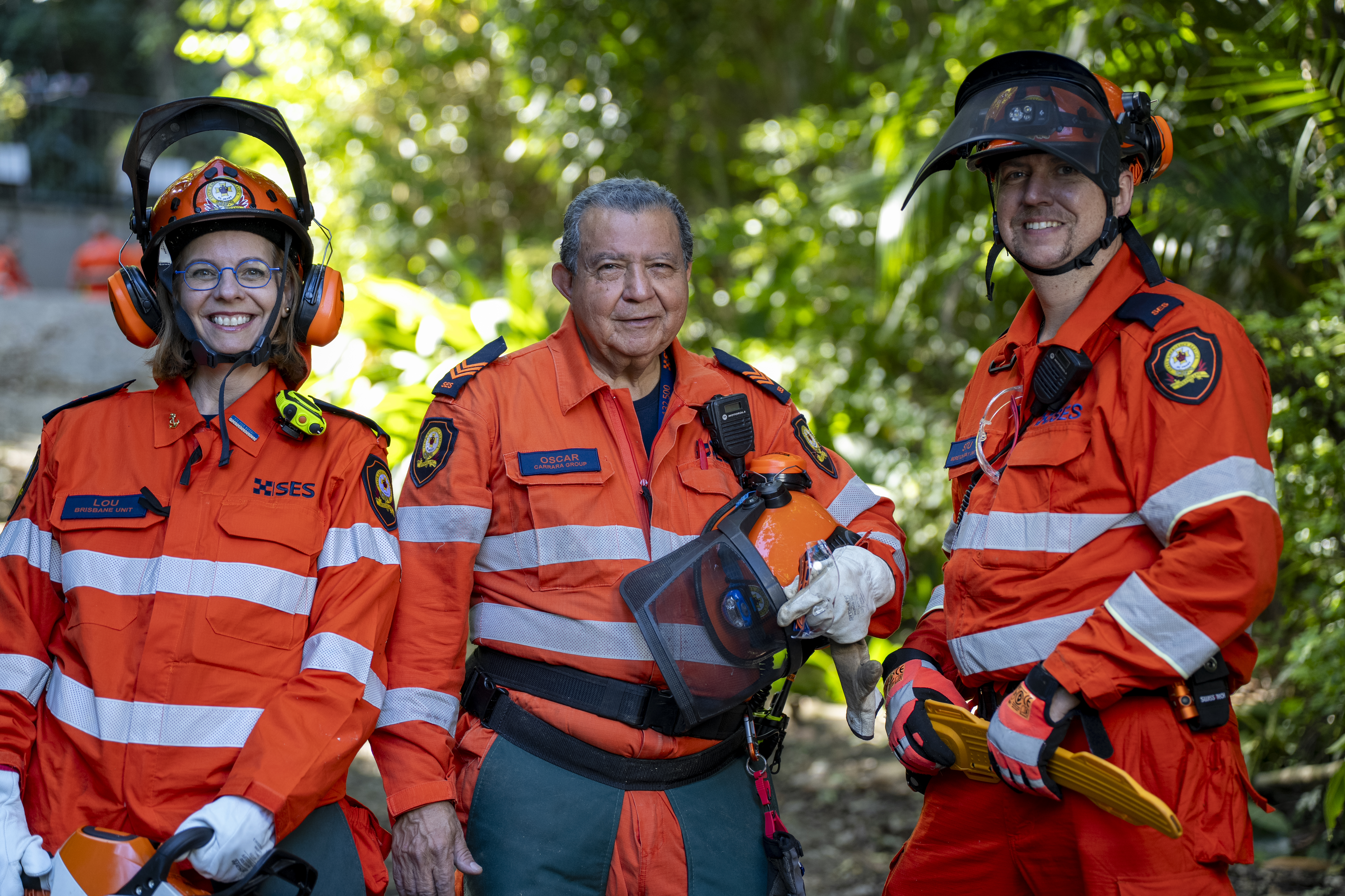 Three SES Volunteers in full protective gear standing in front of tall hedges. They have smiling faces.